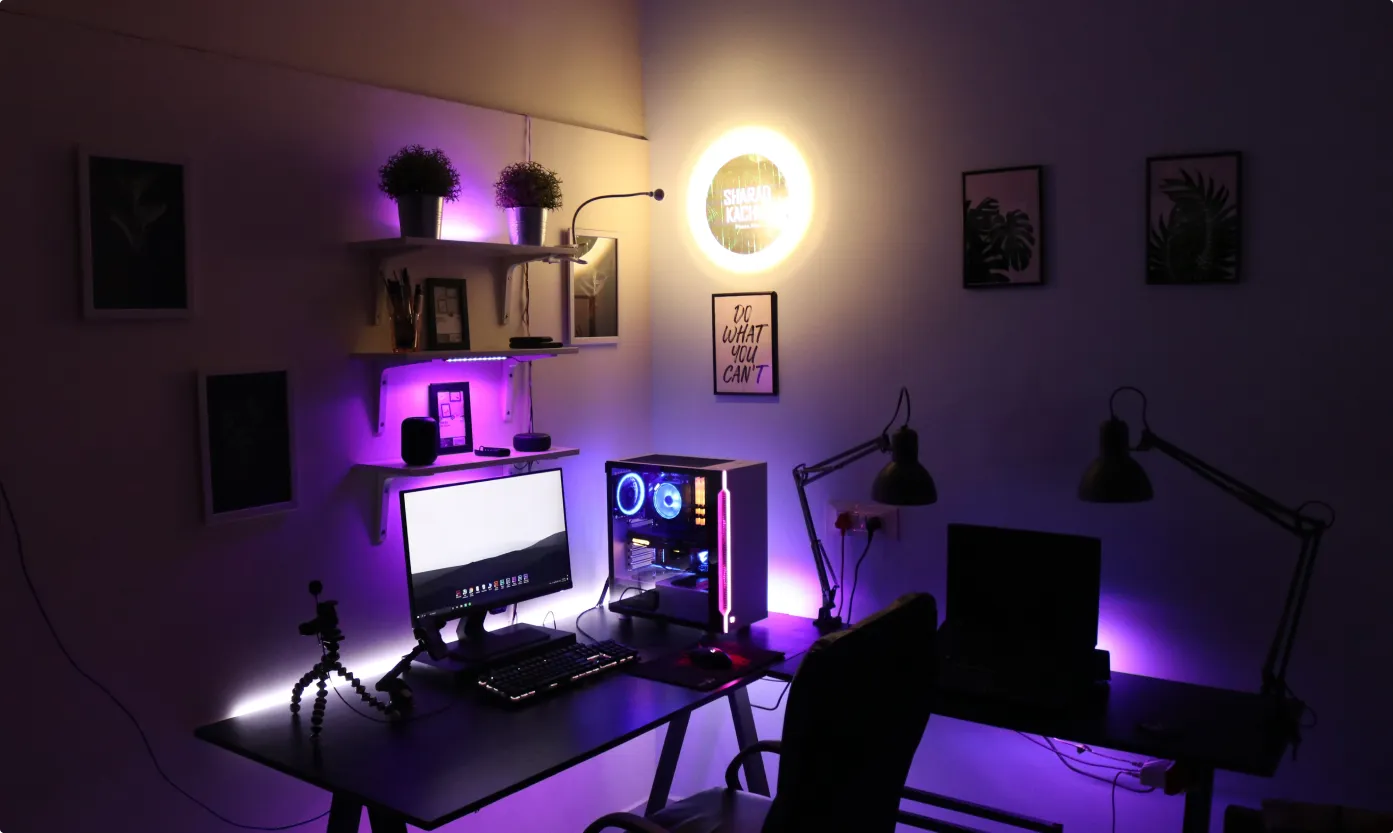Dark gaming desk setup with RGB lighting, computer monitor, PC tower with glowing fans, dual desk lamps, and purple LED accents.